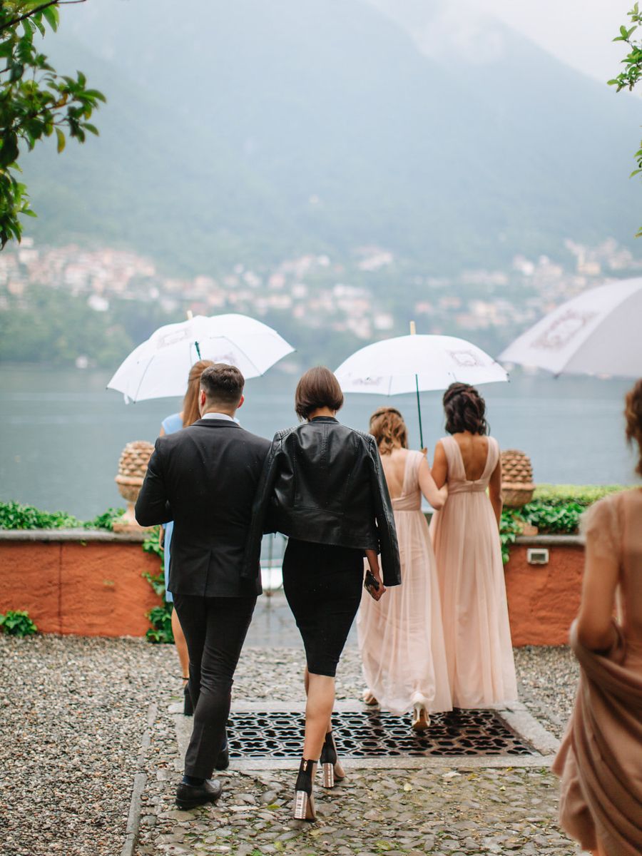 Wedding party stands in a row overlooking Lake Como on a misty day, with umbrellas shielding them from rain.