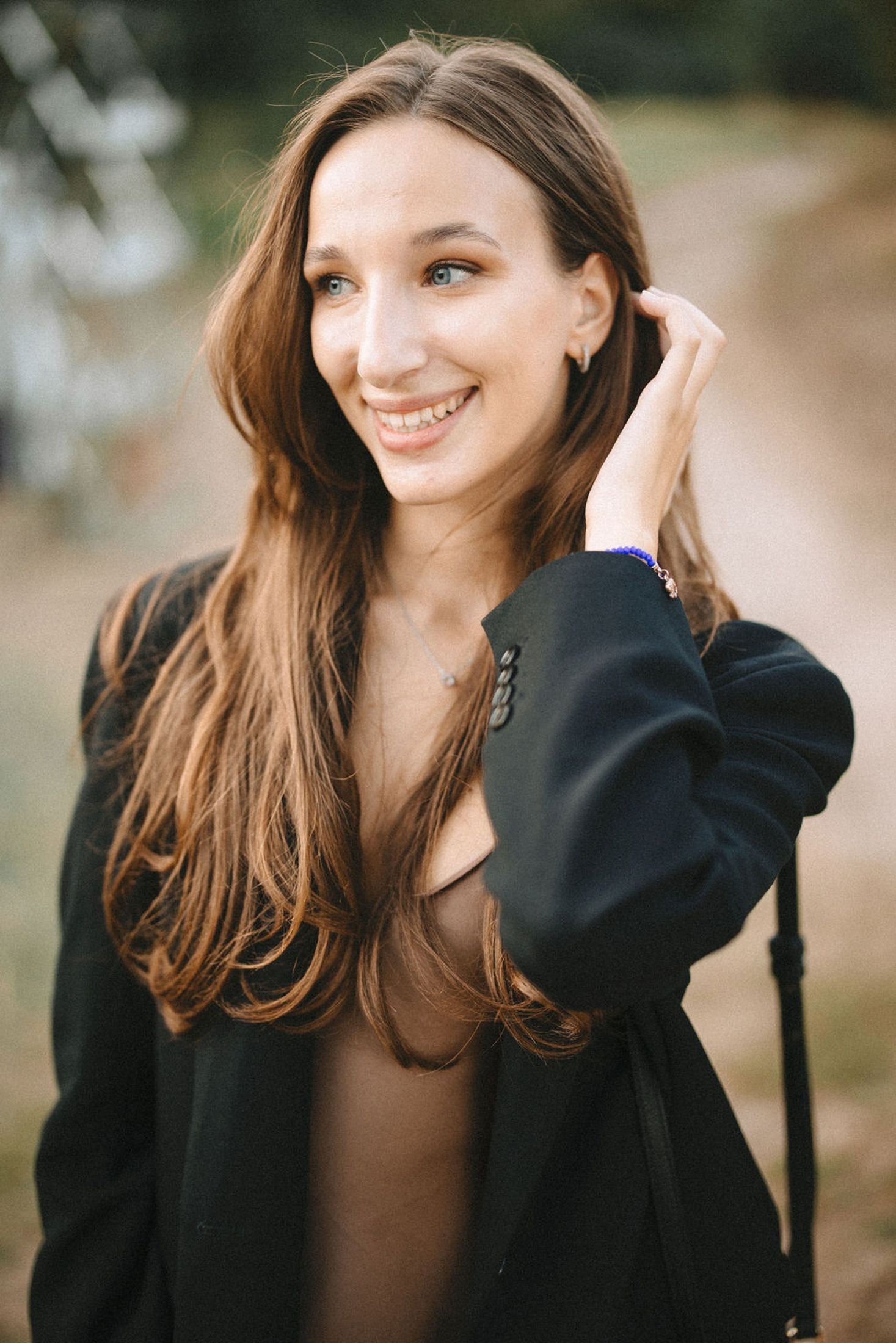 A smiling person with long brown hair wearing a black jacket poses outdoors in soft natural lighting.