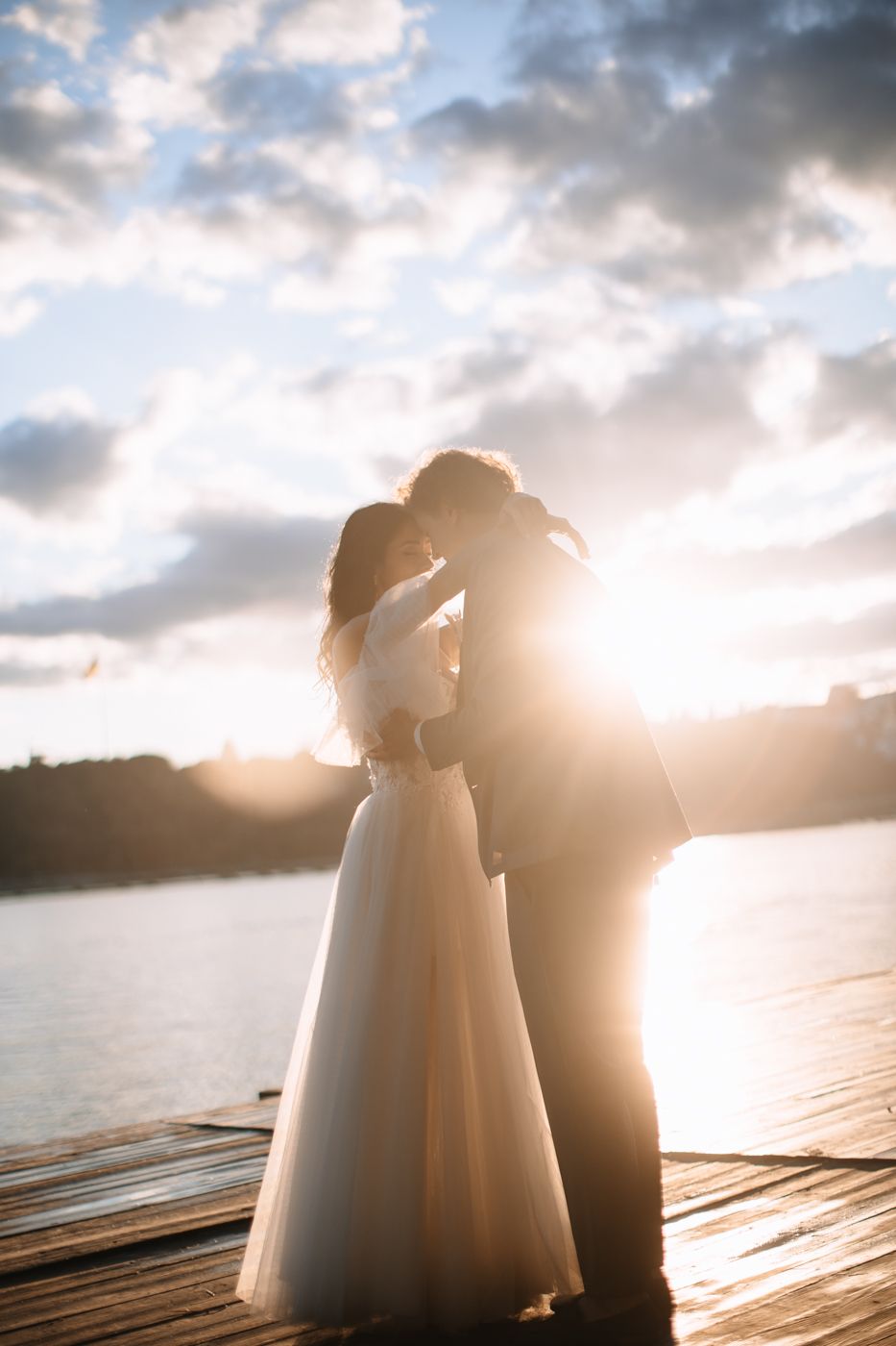 Silhouetted couple embraces on a wooden dock at sunset with dramatic clouds and golden light over the water.