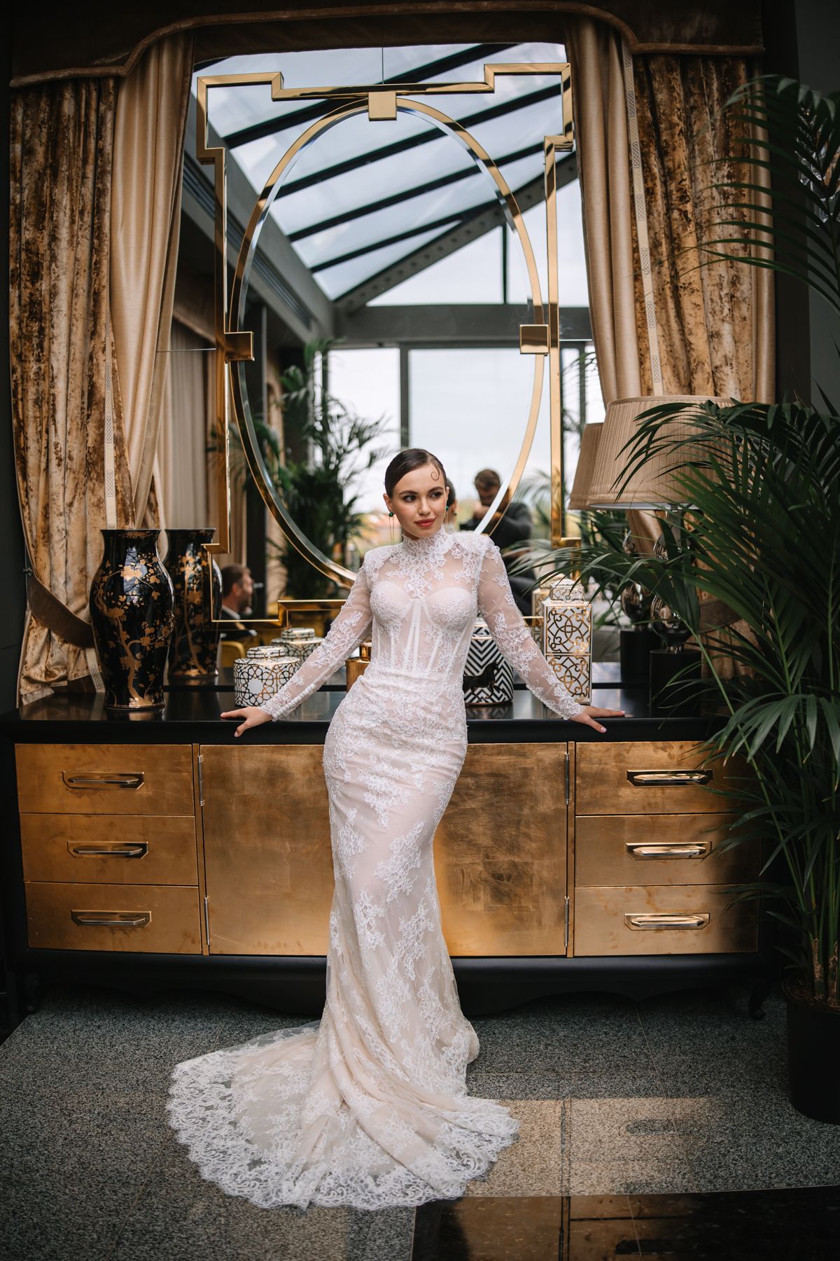 A bride in a lace wedding gown poses in front of wooden cabinets and windows in an elegant indoor setting.
