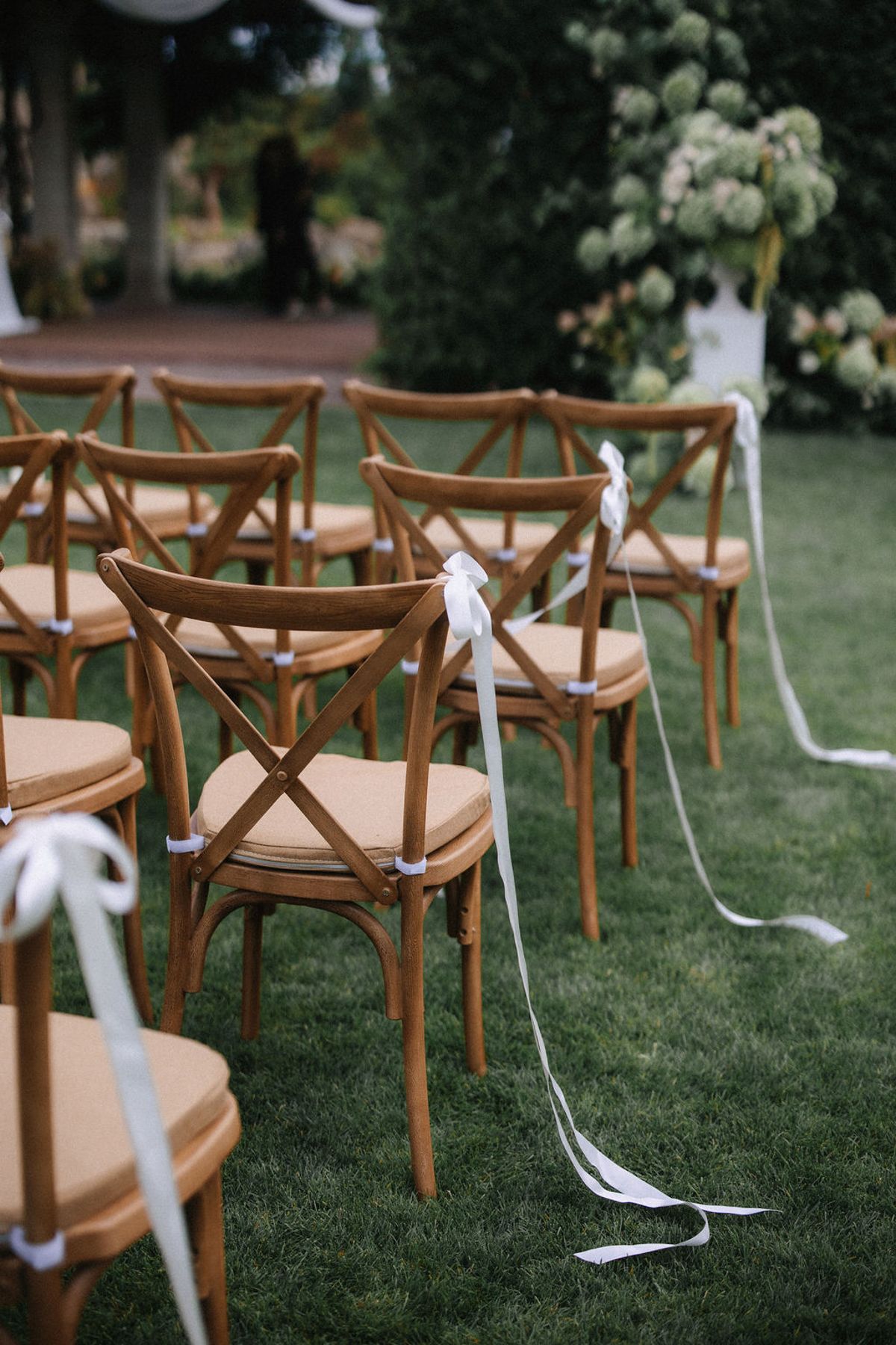Rustic wooden crossback chairs with white ribbons arranged on green grass for an outdoor wedding ceremony.