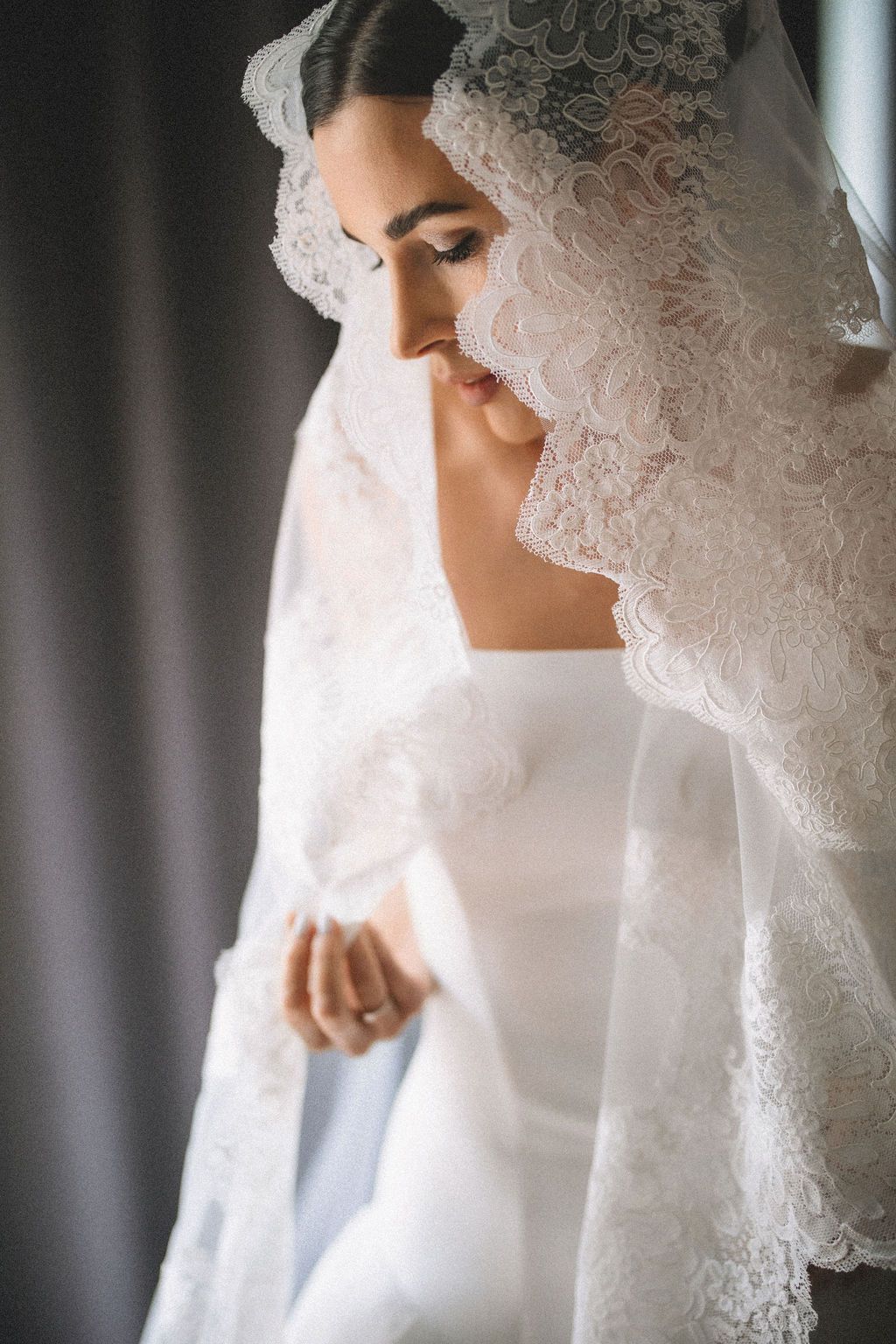 A bride in a white wedding dress and delicate lace veil stands in soft window light with eyes closed.