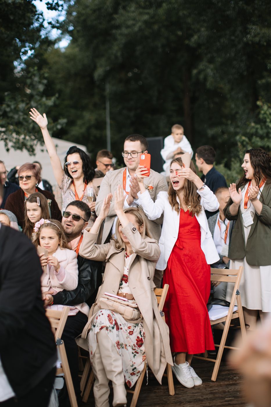 Wedding guests react with joy and excitement during an outdoor celebration, raising their hands in enthusiastic applause.