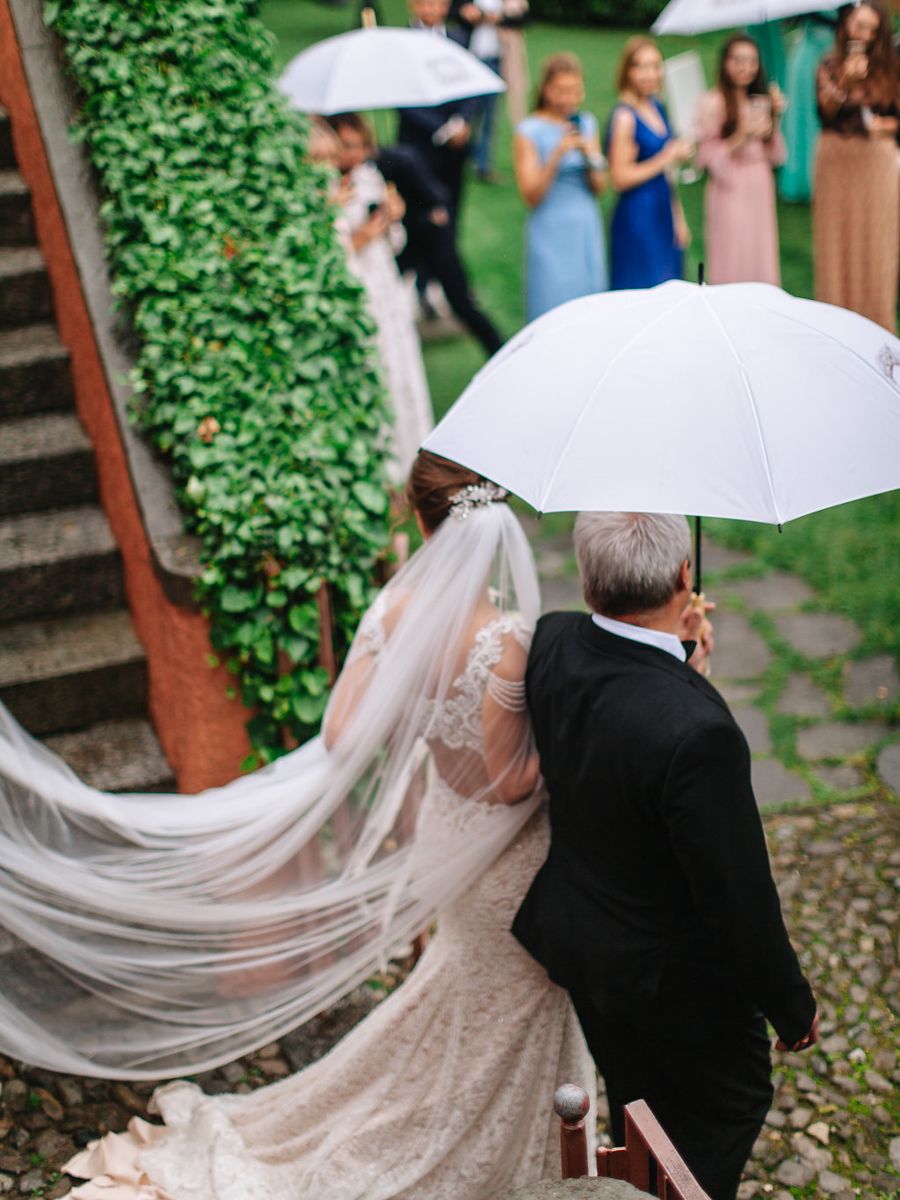 A bride in a lace gown with flowing veil walks up stone steps while a groom in black suit holds a white umbrella.