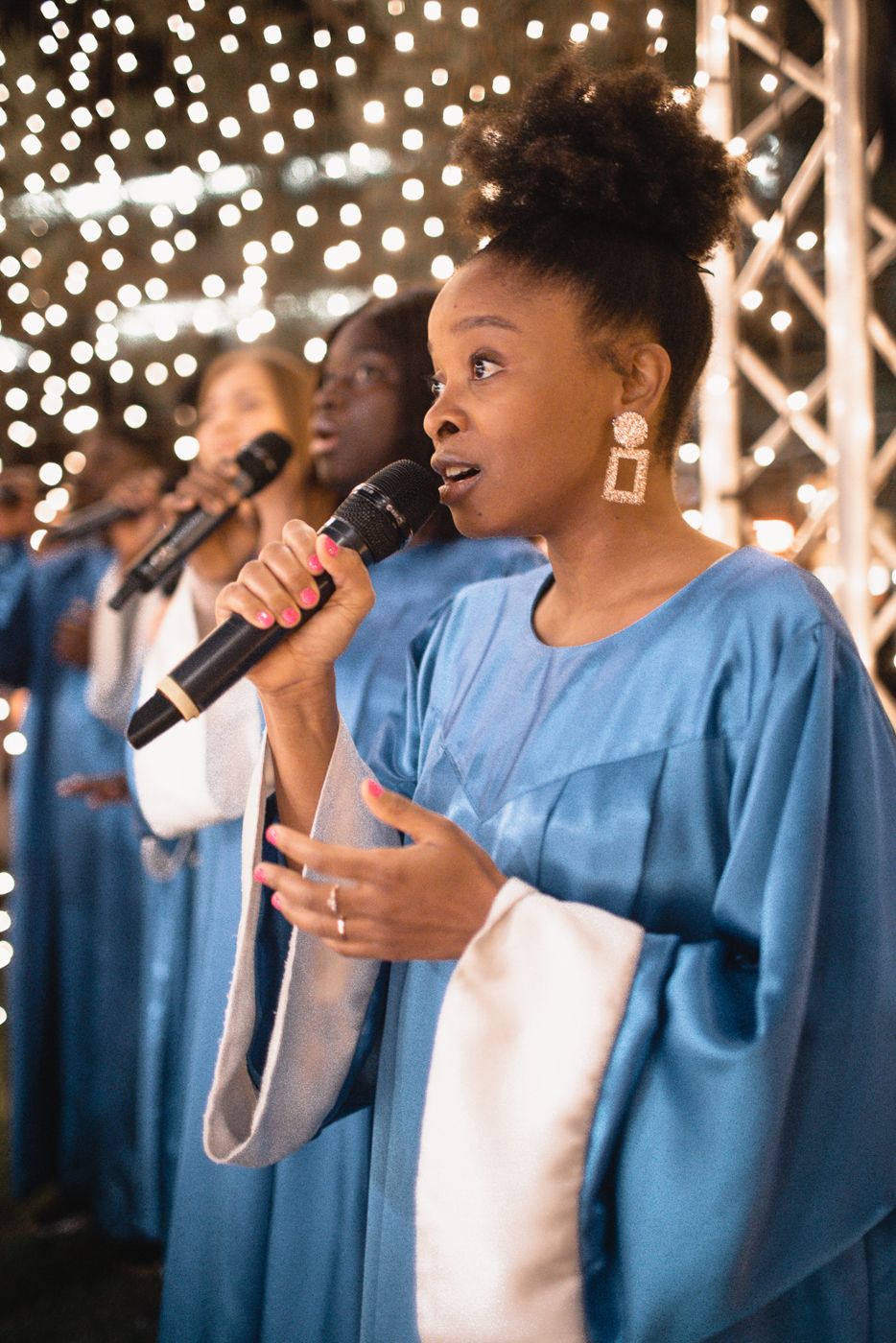 A choir member in blue robes sings into a microphone against a backdrop of sparkling lights.