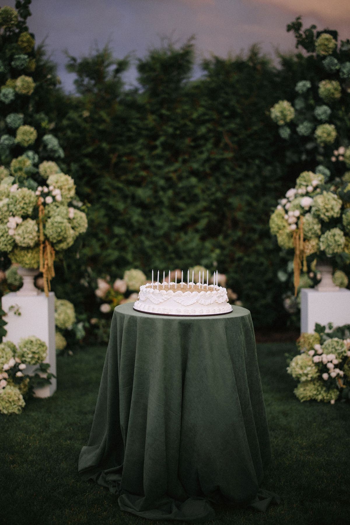 Elegant birthday cake with candles sits on dark green tablecloth against lush garden backdrop at twilight.
