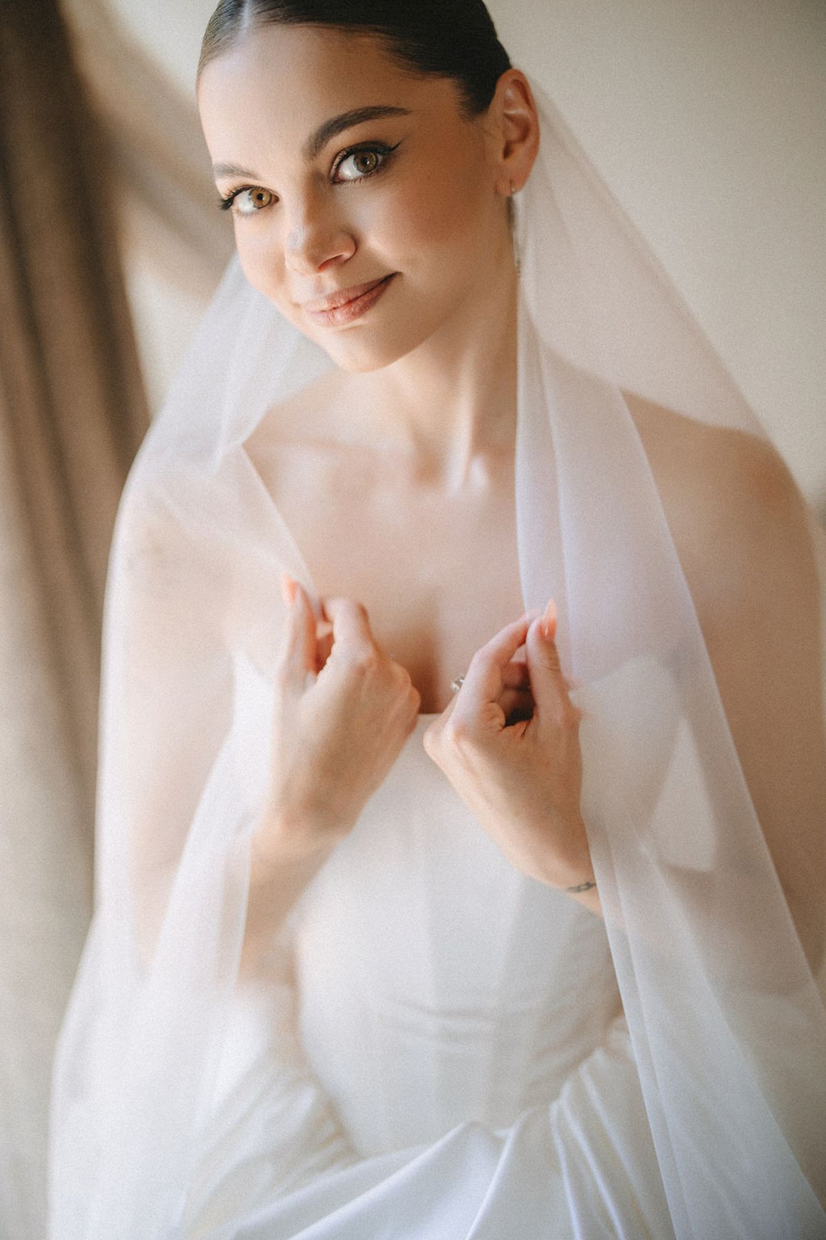 Elegant bride in white strapless gown adjusts delicate veil near window with soft lighting.