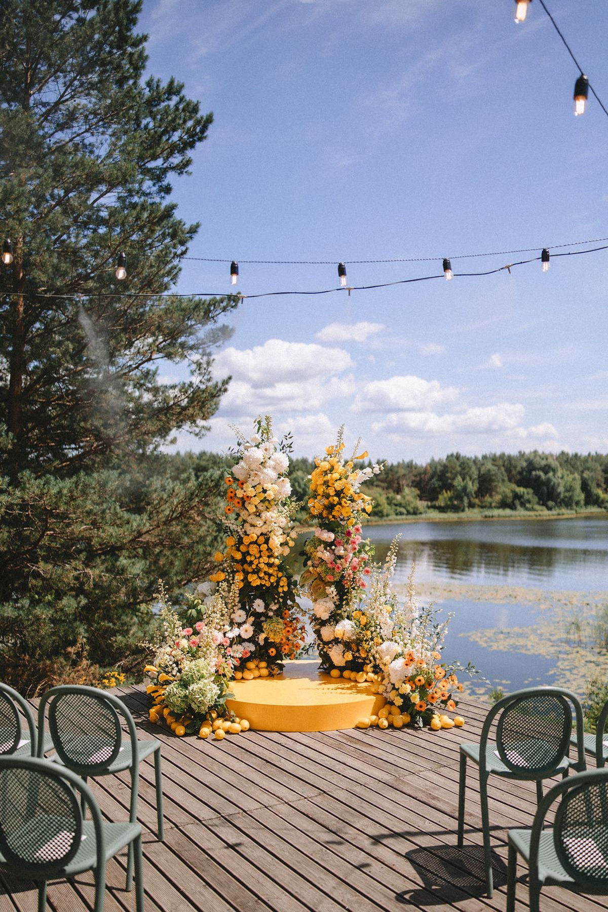 Wooden deck overlooking a lake with green chairs and orange floral arrangements under string lights on a sunny day.