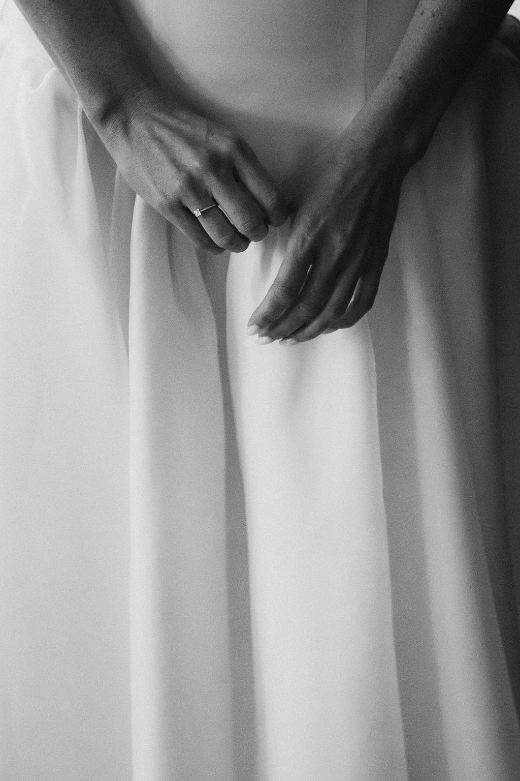 Hands gently grasp the folds of a flowing white dress in an intimate black and white photograph.