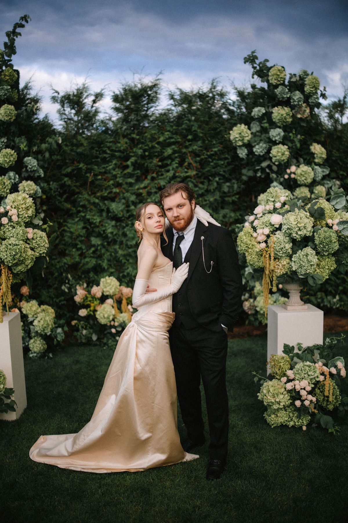 A couple in formal wedding attire pose together in front of a lush green hedge with white hydrangea floral decorations.