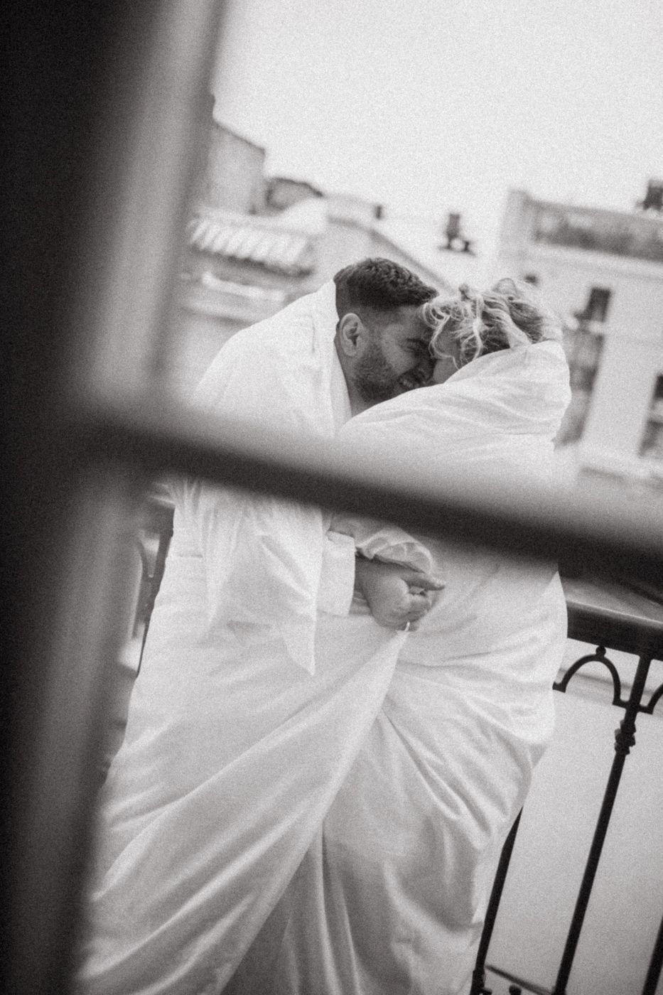 Black and white photo of couple in white robes embracing on balcony.