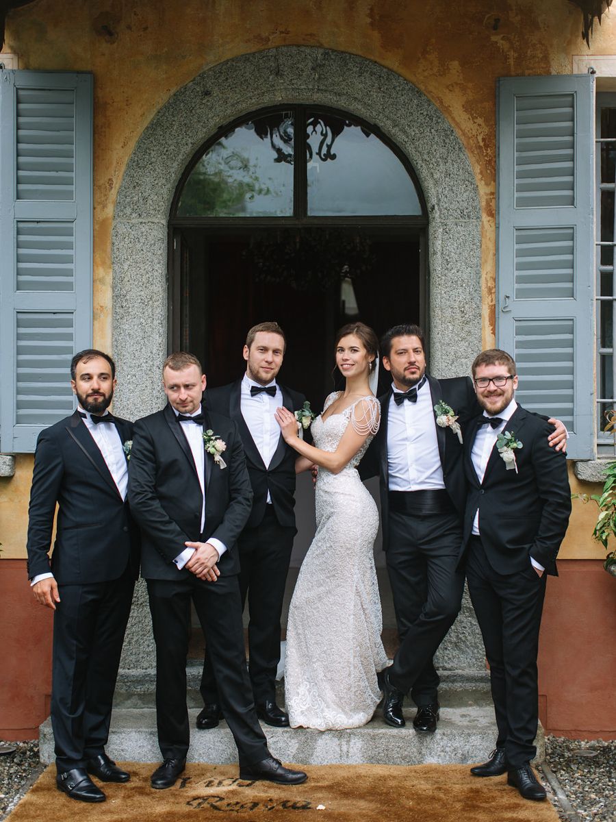 Wedding party in formal attire poses playfully in front of an elegant Mediterranean-style building with arched windows.