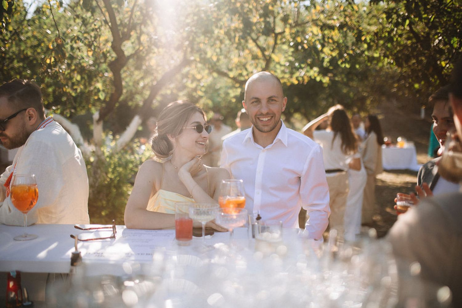 People enjoying drinks and conversation at an outdoor garden party with golden sunlight filtering through trees.