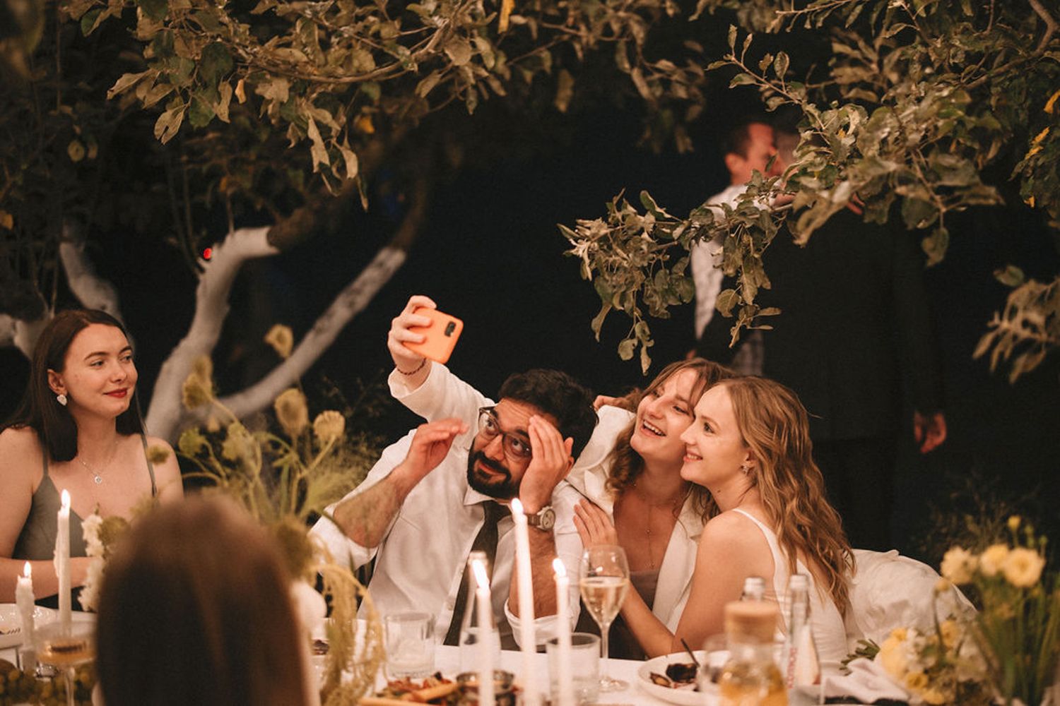 Wedding guests laugh and celebrate together at an outdoor reception dinner table under string lights and trees.