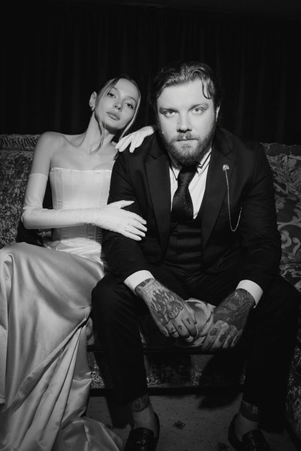 A black and white portrait of a couple seated together on a couch, with her in a white gown and him in a dark suit.