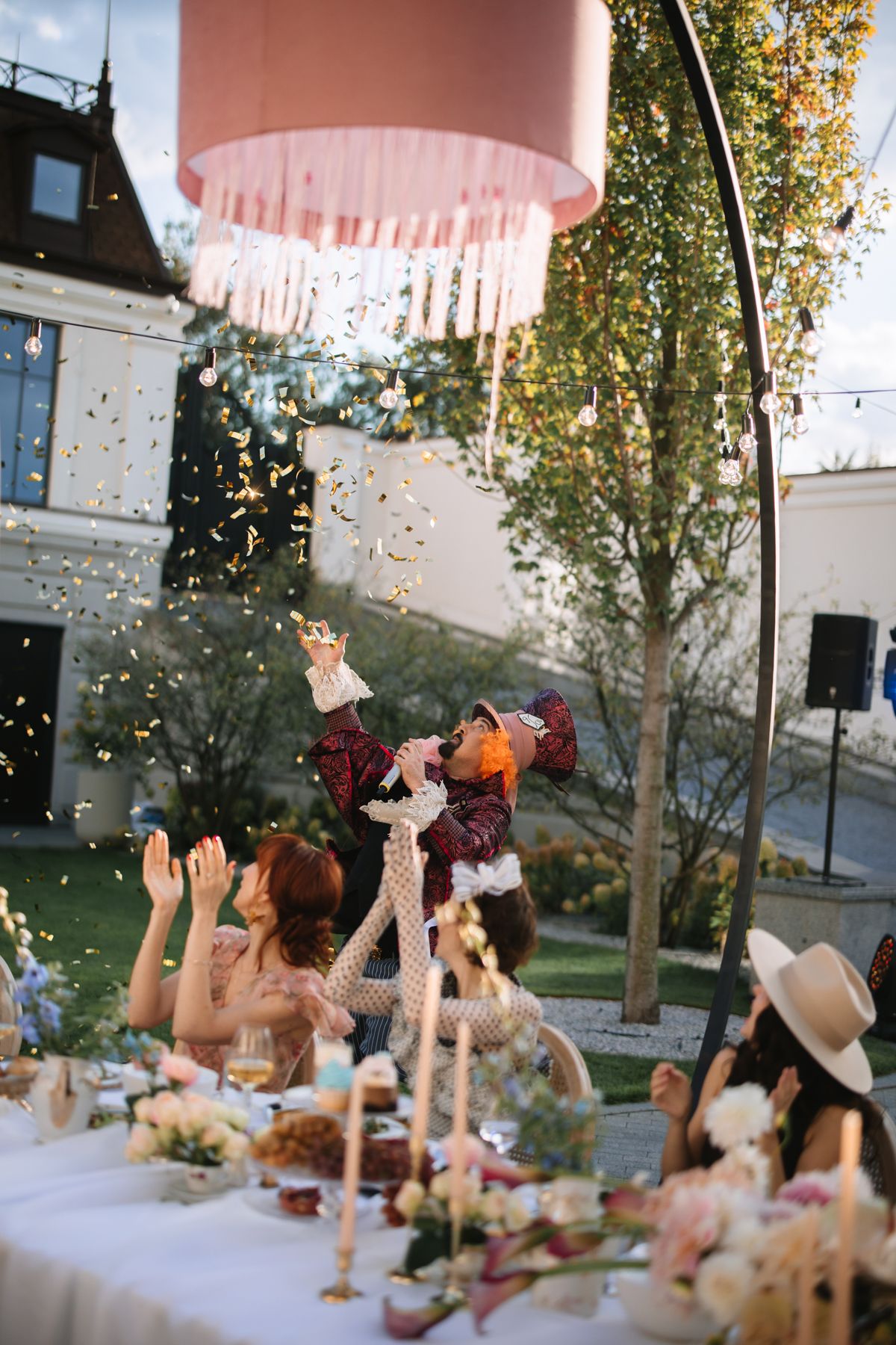 Wedding guests reach up to catch falling confetti beneath a pink chandelier at an outdoor reception celebration.