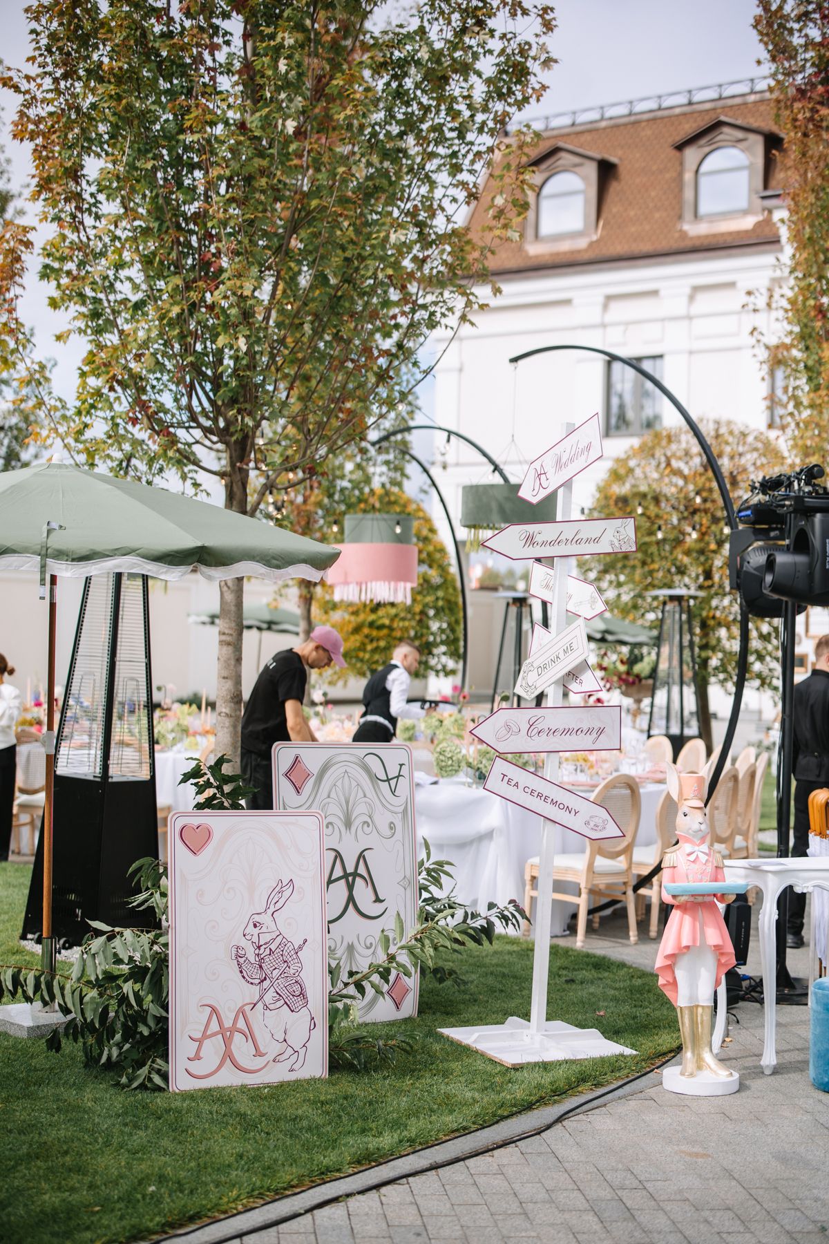 Outdoor event setup with white umbrellas, decorative panels, and pink accents along a garden pathway.