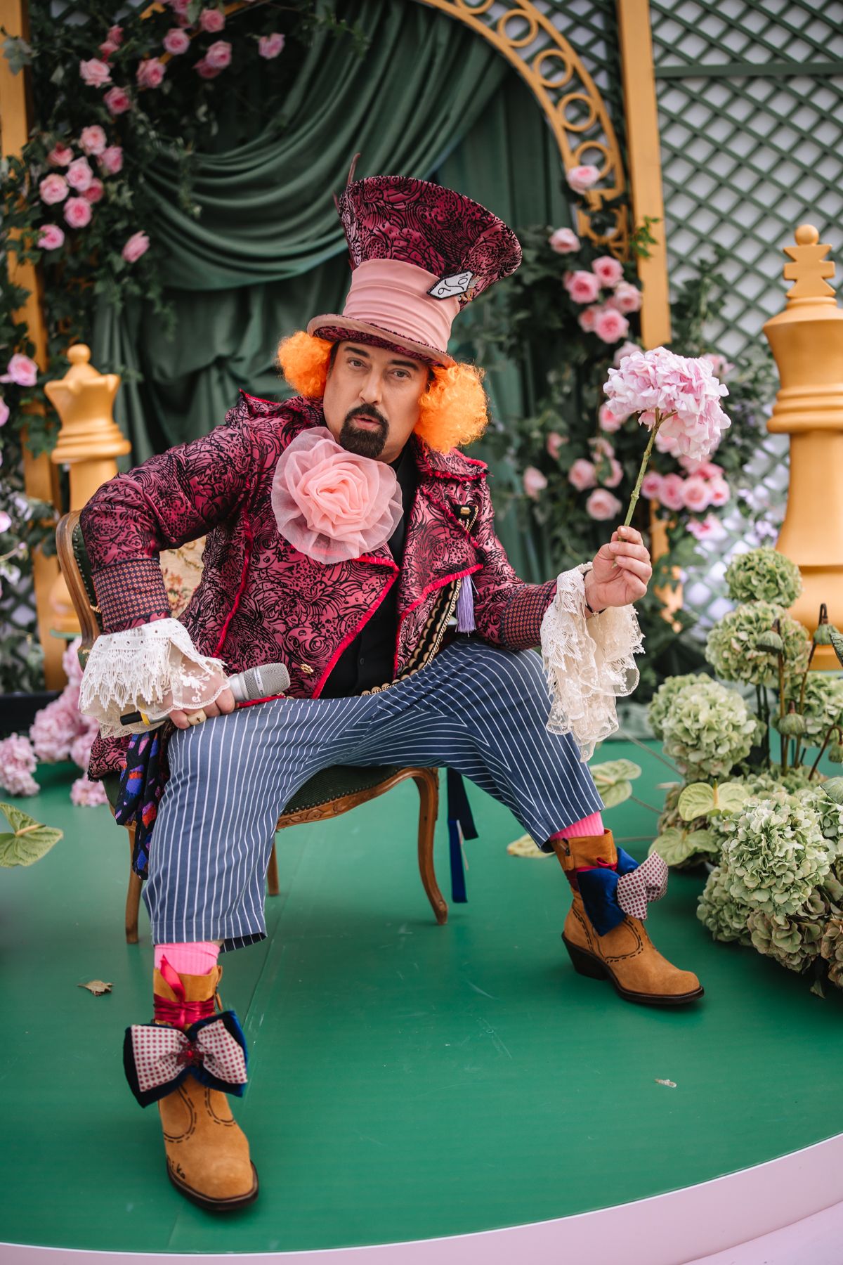 Performer in burgundy Victorian costume and hat strikes playful poses on green stage with pink floral decorations.