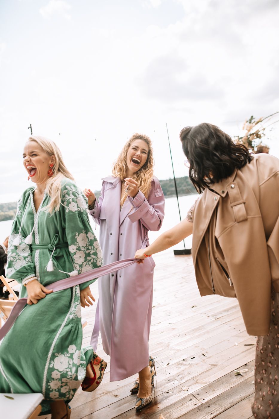 A group of friends in pastel outfits laugh and dance together on a wooden deck by the water.