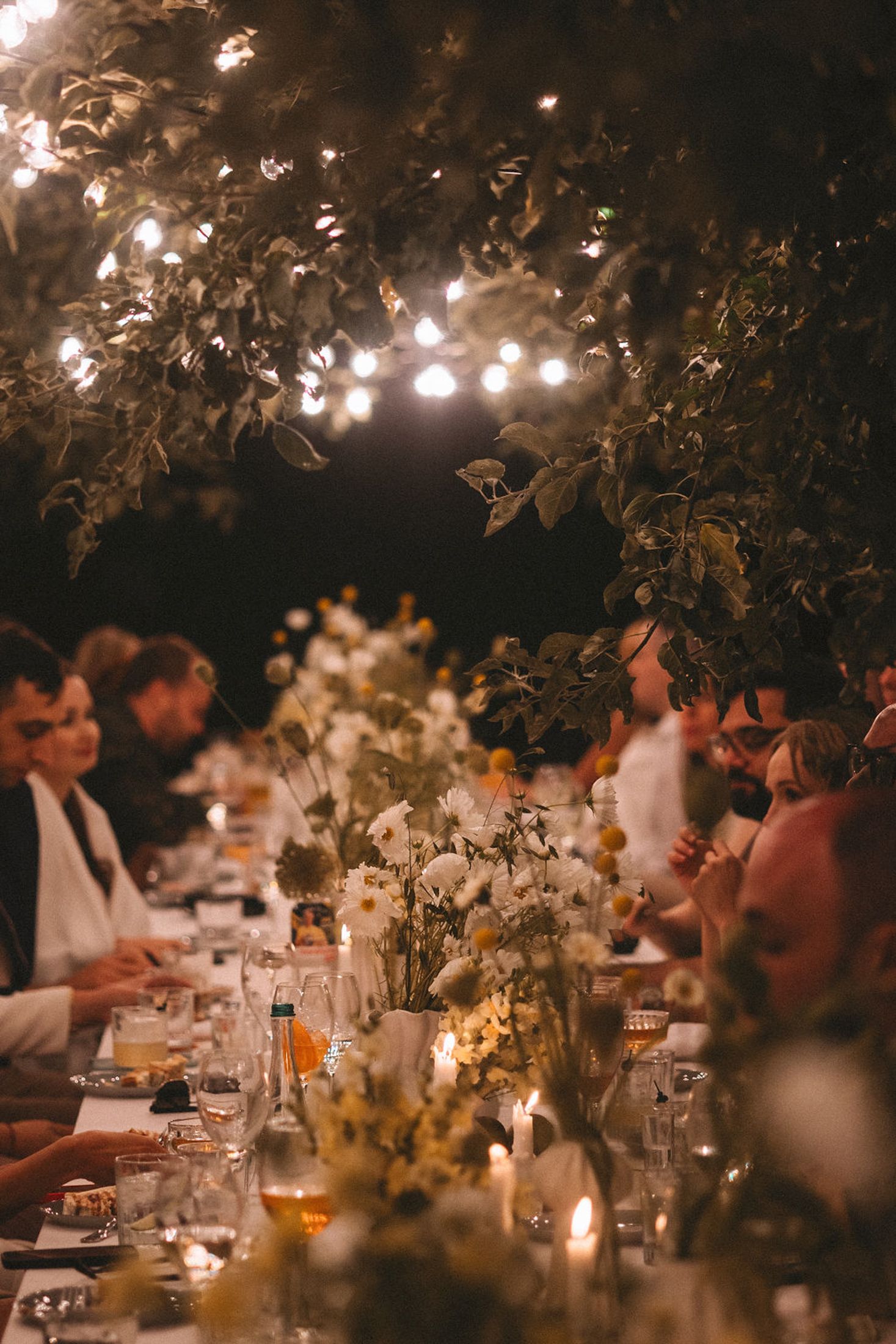Romantic outdoor dinner party at dusk with glowing candles and floral centerpieces on long tables under trees.
