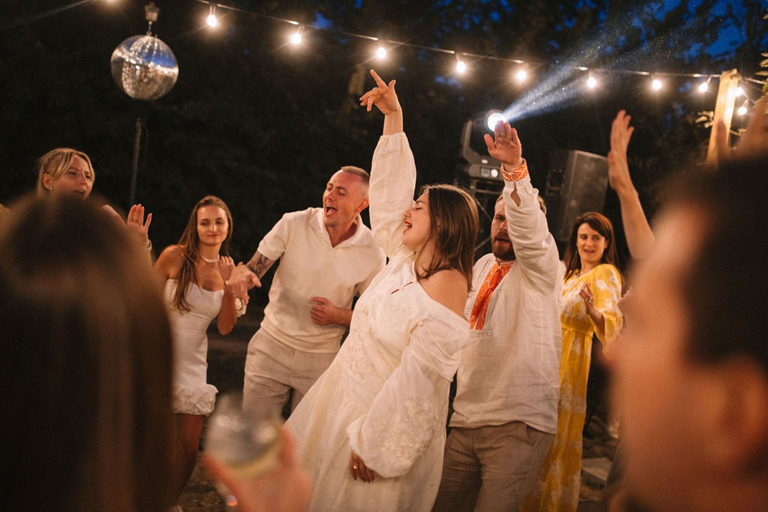 Wedding guests in white attire dance and celebrate under string lights at an outdoor evening reception.