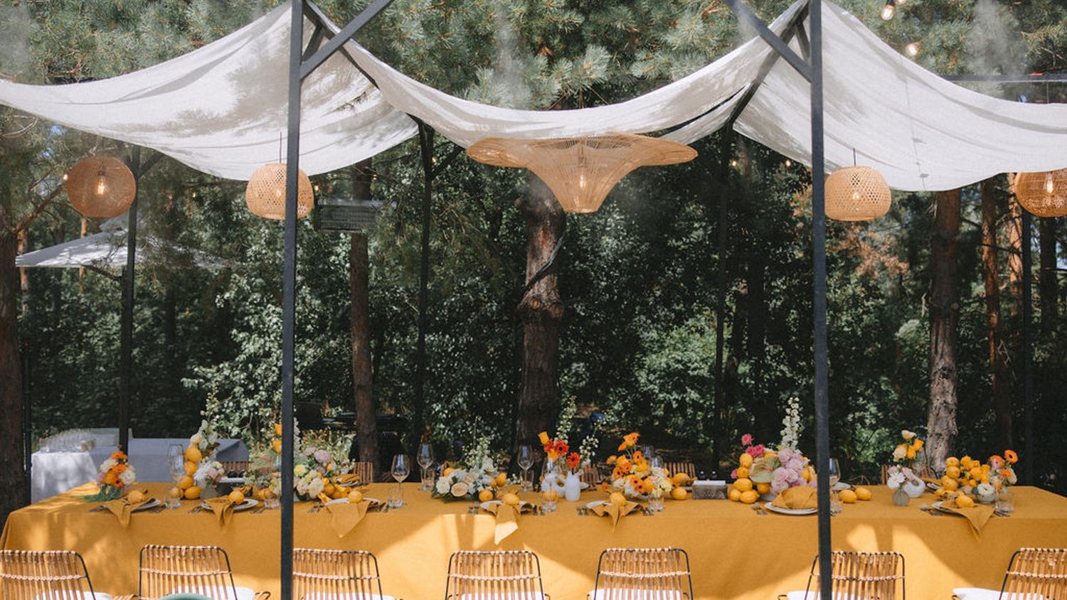 Outdoor wedding reception setup with yellow tablecloths and draped white canopies in a wooded setting.