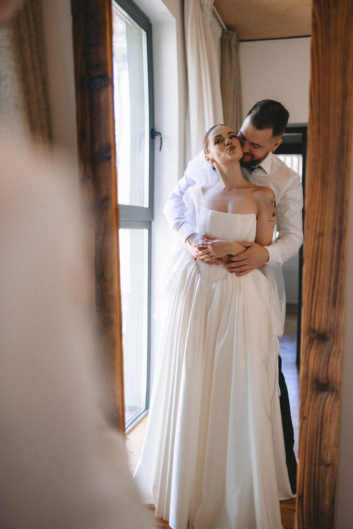 Romantic wedding moment of couple embracing by window in rustic interior with flowing white dress and natural light.