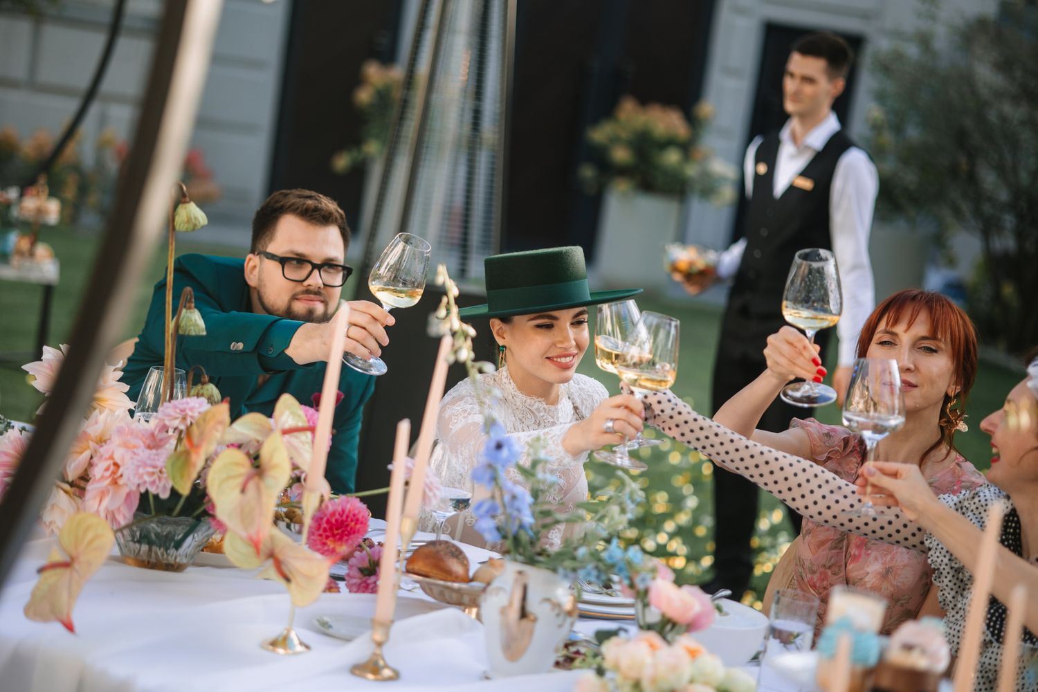 People raise wine glasses in a toast at an outdoor celebration with floral table decorations.