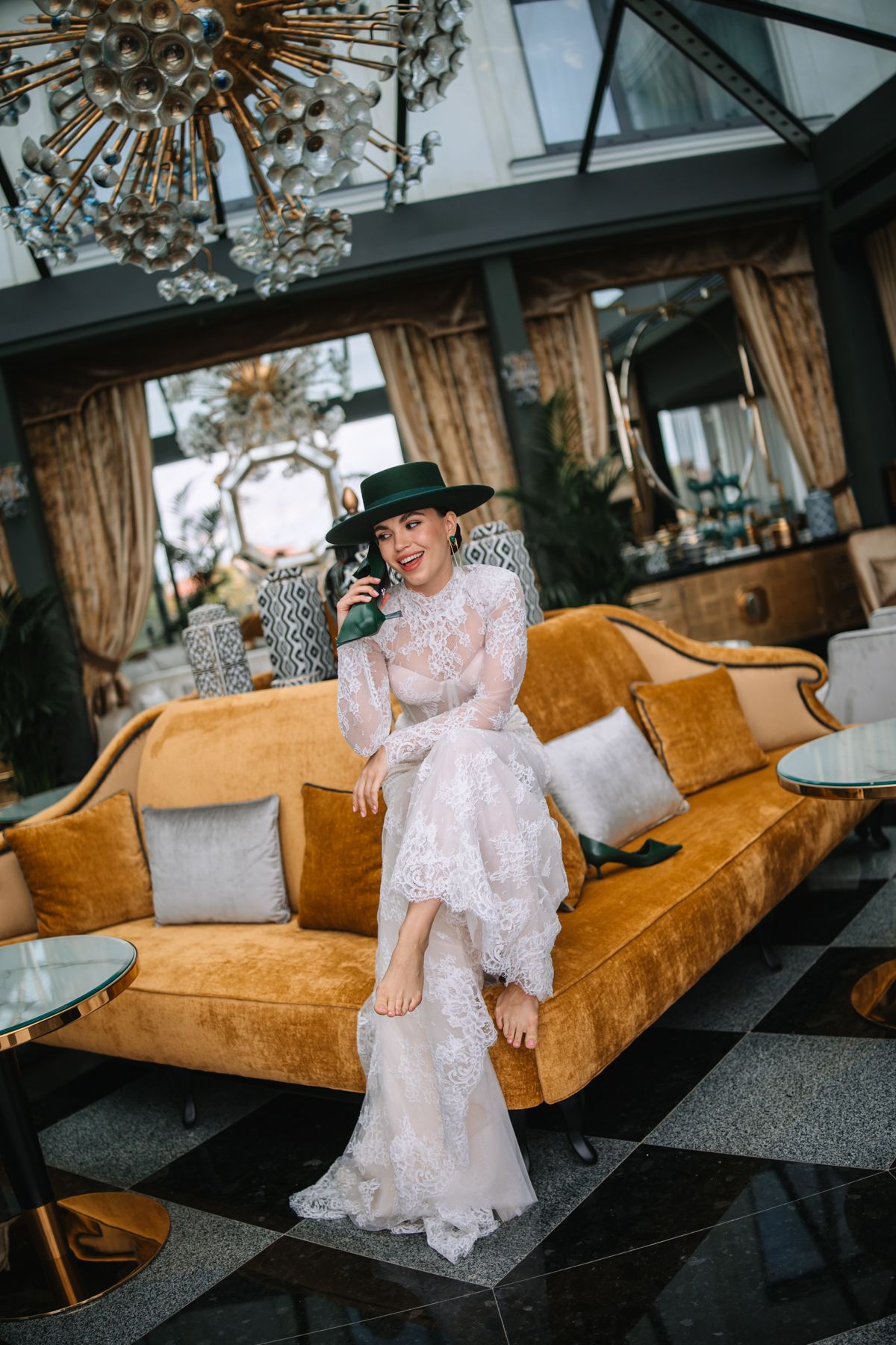 Elegant figure in white lace dress and black hat sits on luxurious orange velvet couch under crystal chandelier.