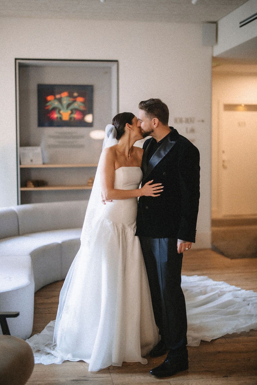 A romantic wedding moment captured indoors with a bride in white gown and groom in black suit sharing a tender kiss.
