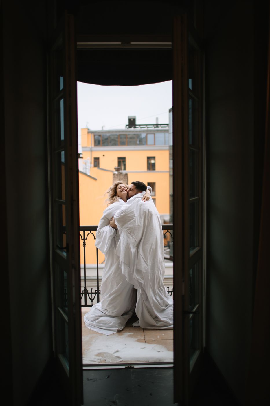 Couple embracing on balcony framed through doorway with urban backdrop.