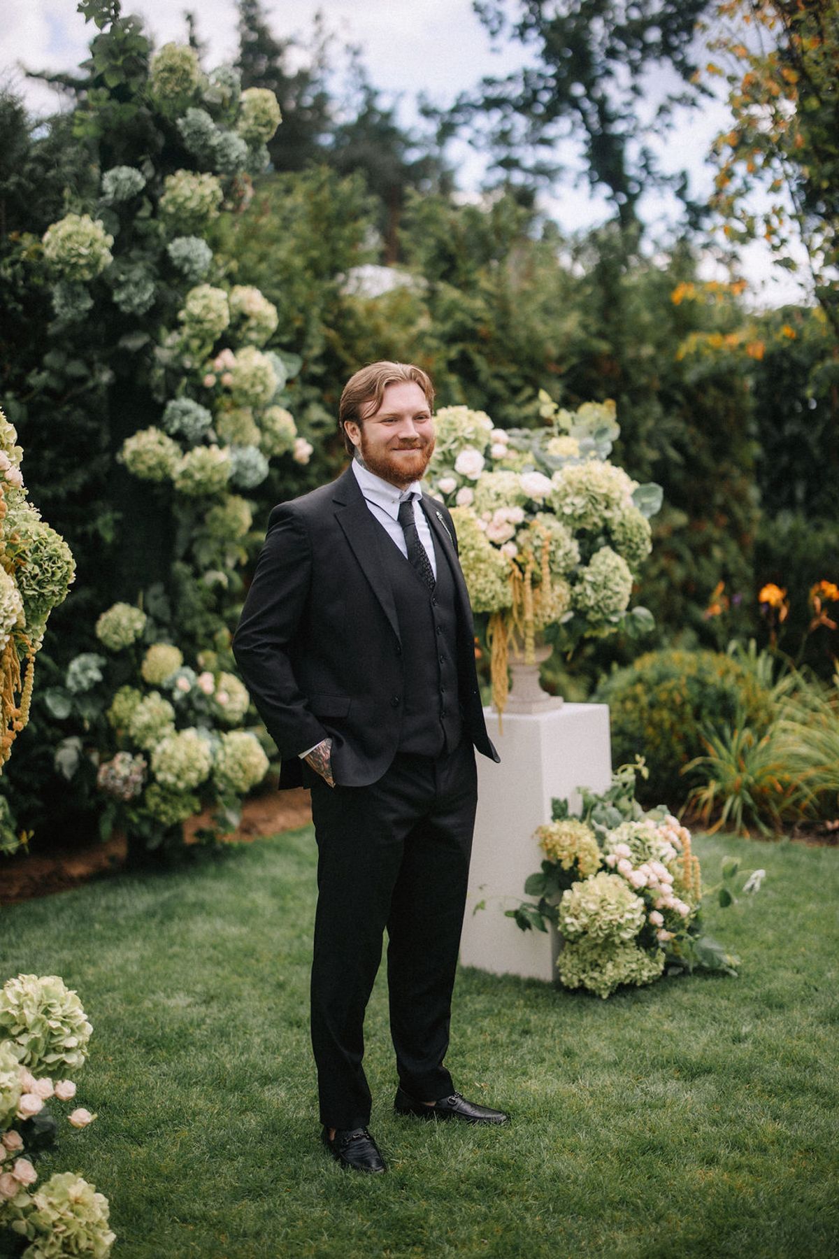 A person in a black suit stands near elegant white hydrangea arrangements in an outdoor garden setting.
