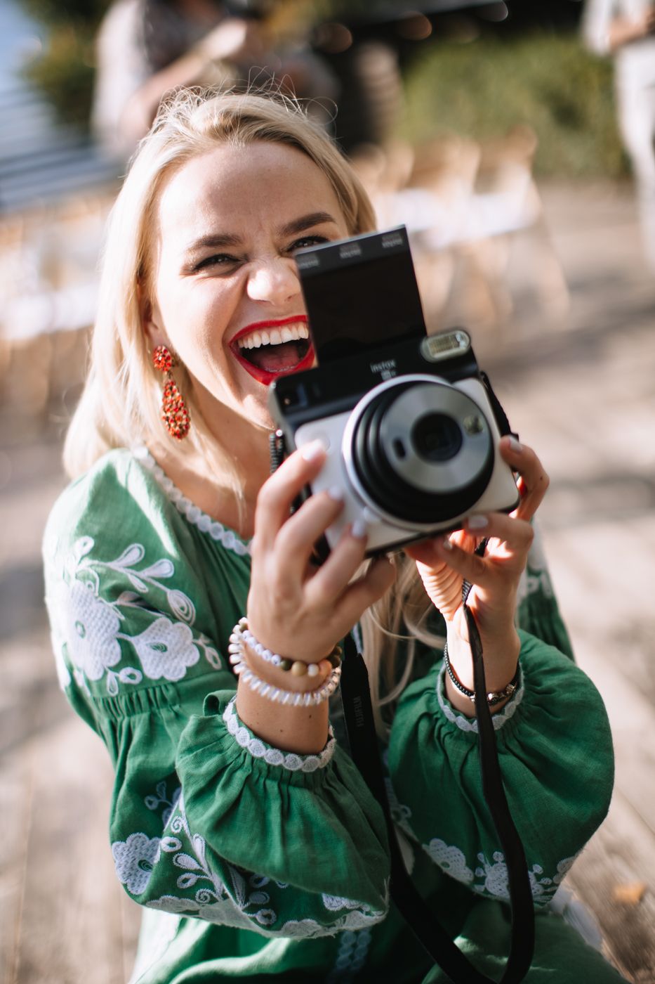 A photographer in a green floral dress and pearl necklace joyfully takes pictures with a vintage camera outdoors.