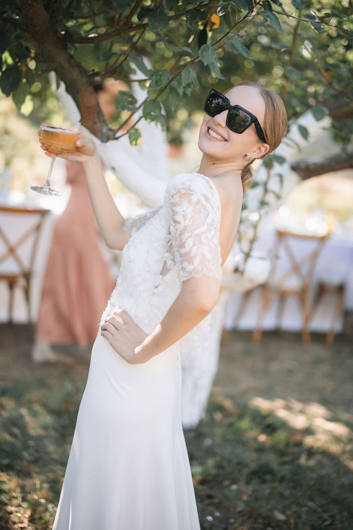 A person in a white lace wedding dress and sunglasses poses playfully at an outdoor garden reception.