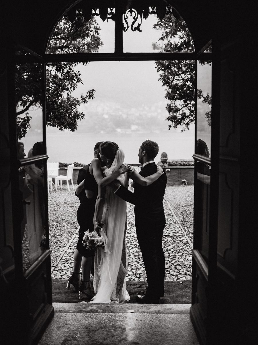 A silhouetted wedding party stands in an open doorway overlooking a scenic waterfront view in black and white.
