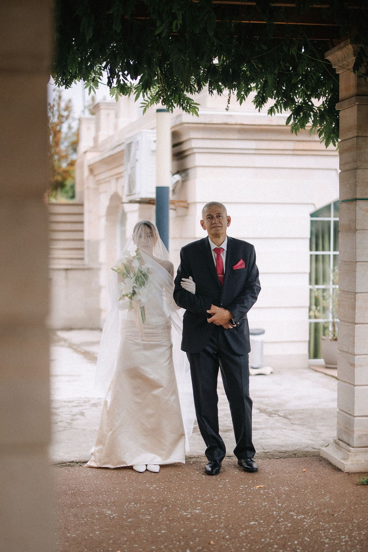 A bride in a white gown and veil stands with an older gentleman in a black suit with red tie under an archway.