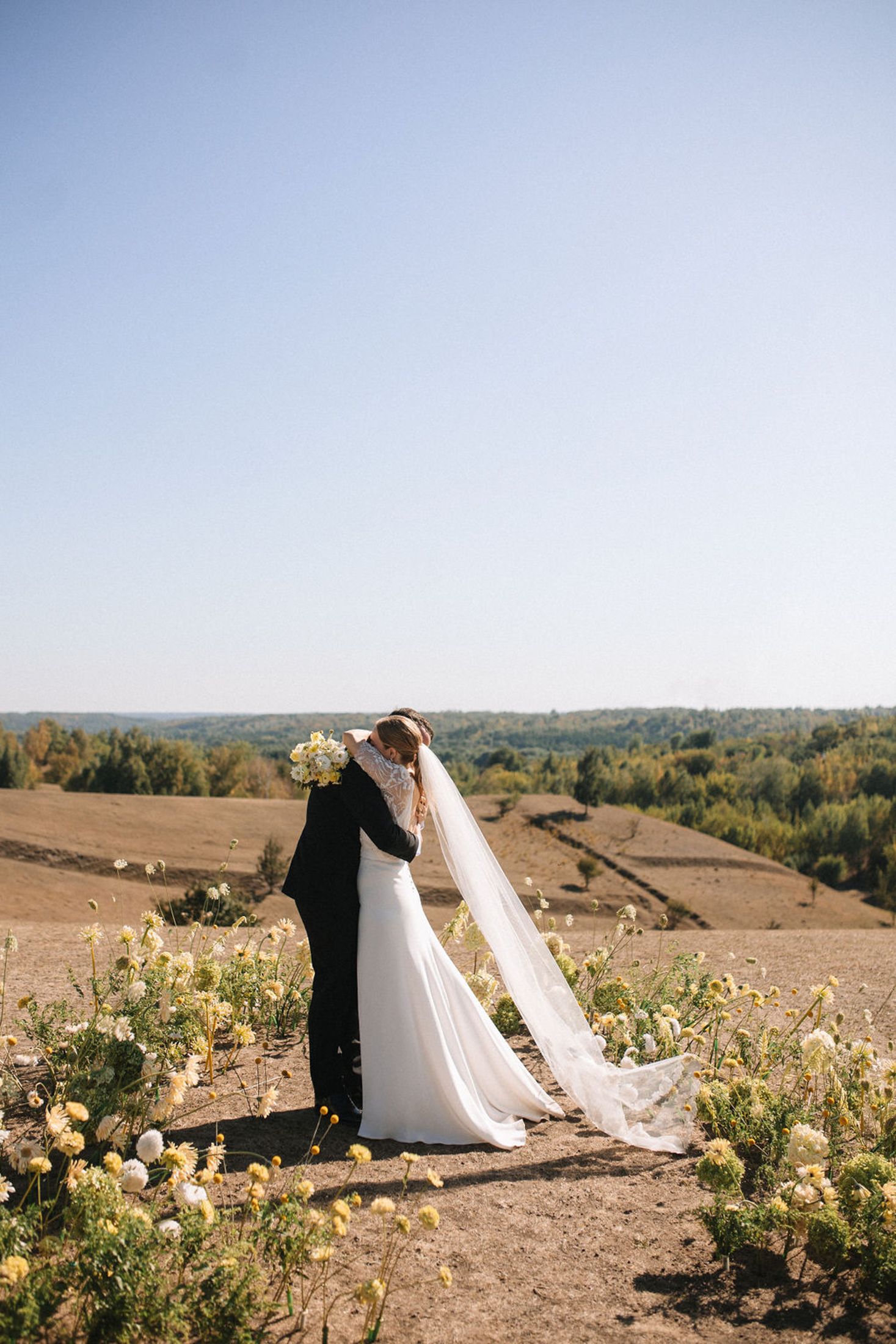 A romantic wedding couple stands on a hilltop overlooking a scenic autumn landscape with wildflowers in bloom.