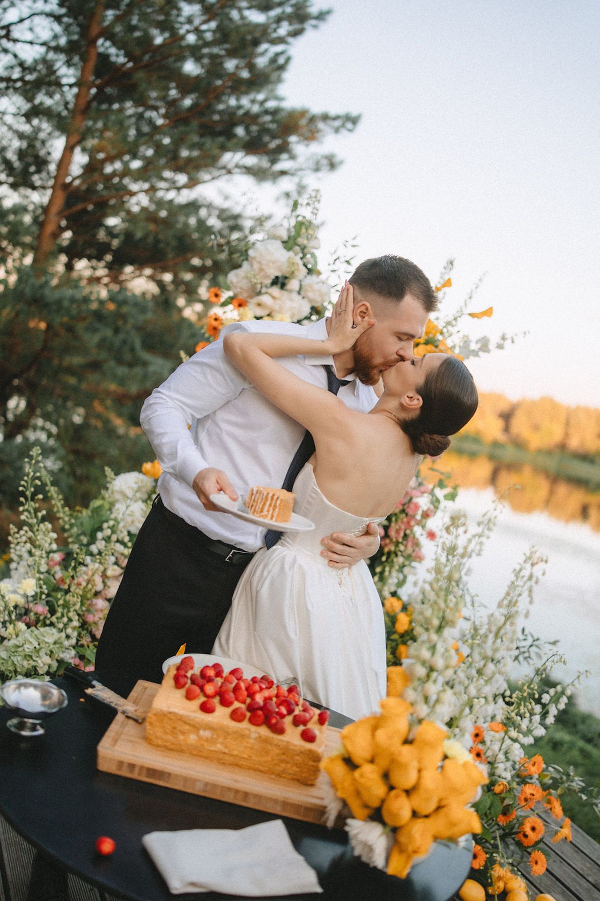 Romantic outdoor picnic scene with fresh fruits and flowers arranged on wooden boards by a lake at sunset.