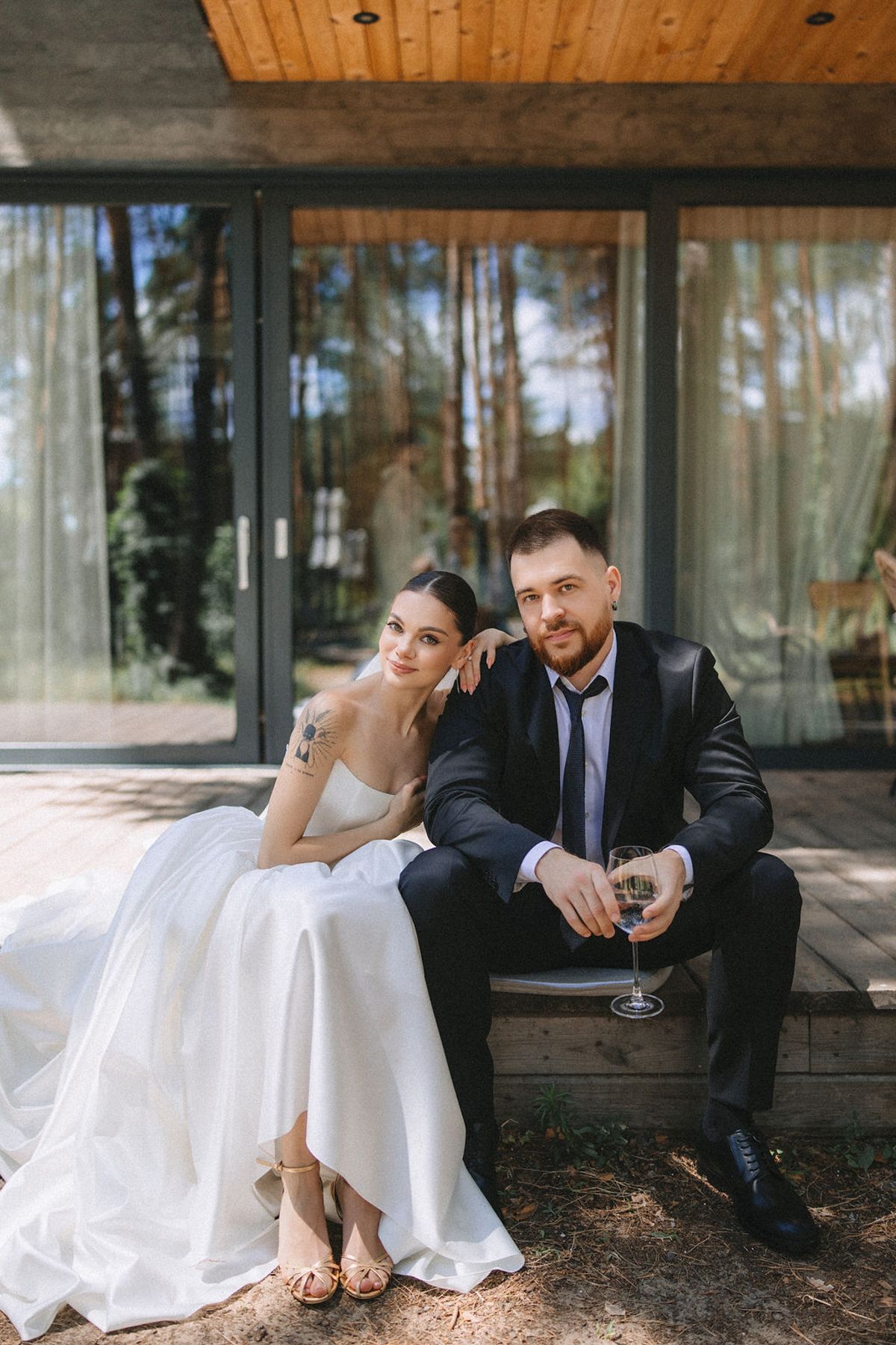 A couple in wedding attire relaxes together on a rustic bench outside a modern building with glass walls.
