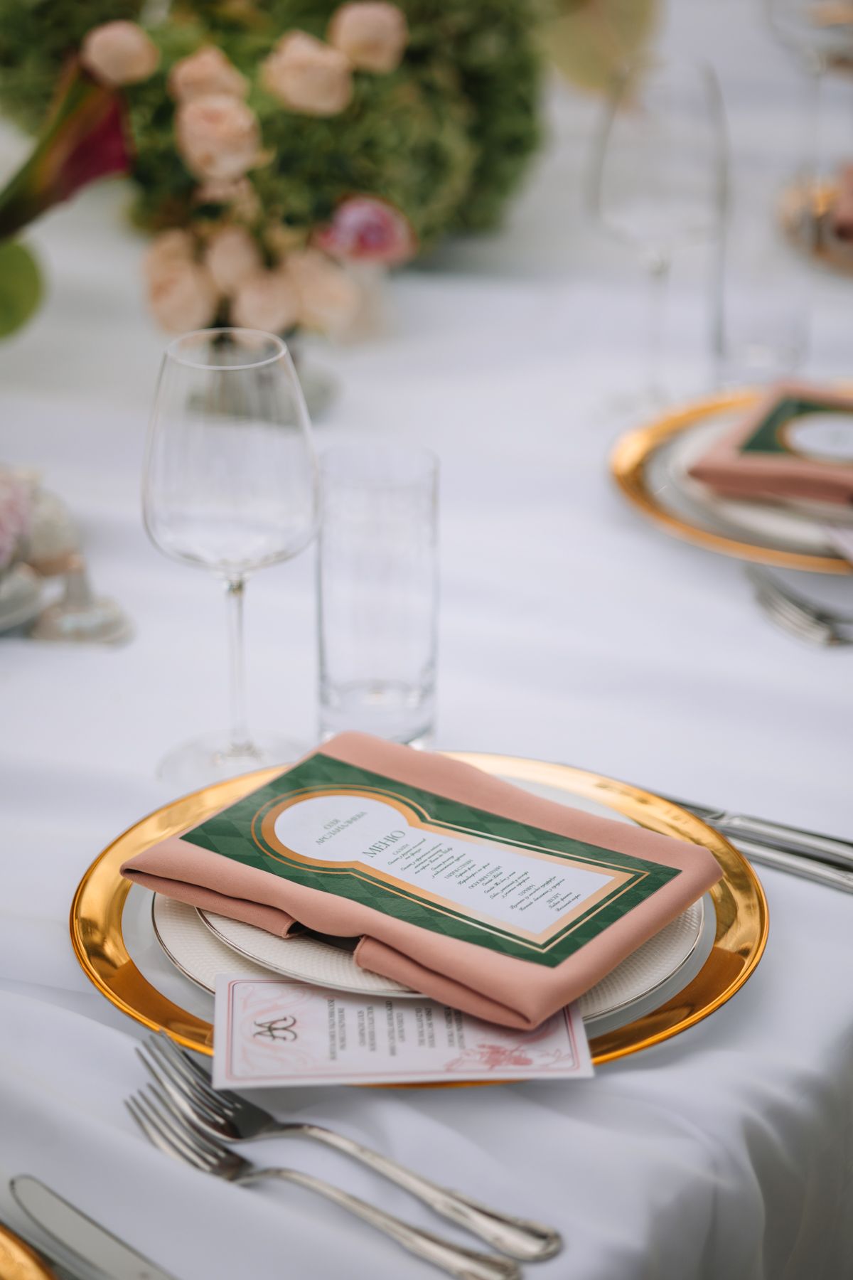 Elegant wedding place setting with gold-rimmed plates, blush pink napkins, and emerald green menu cards on white tablecloth.