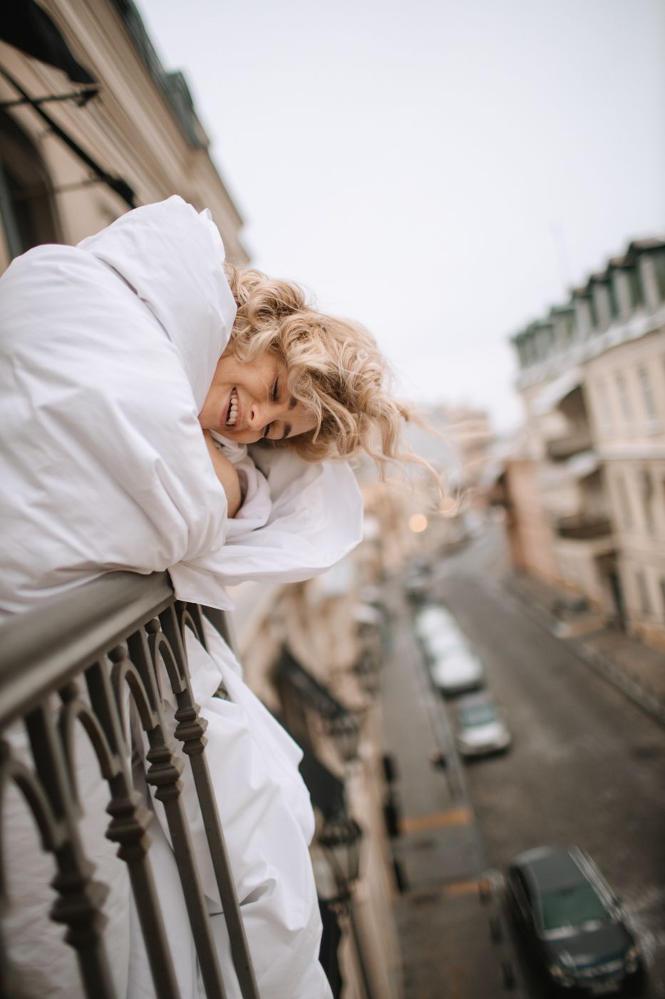 Person in white robe leans over balcony railing with curly blonde hair flowing in urban setting.
