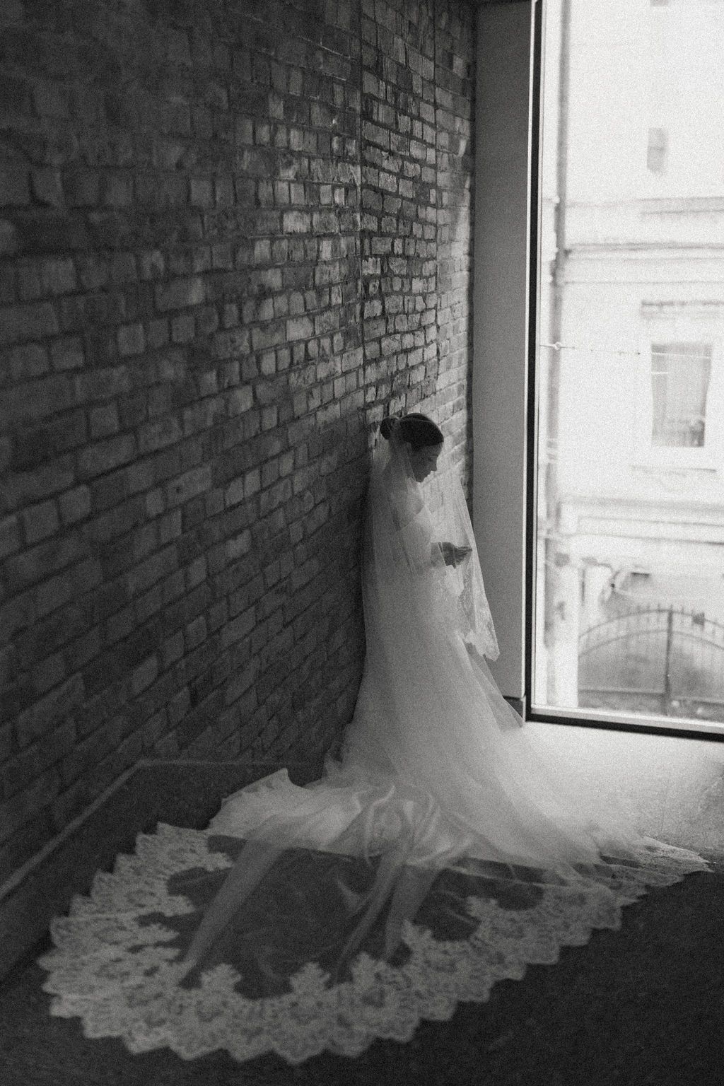 A black and white series showing a wedding dress with a flowing train displayed against a brick wall near a window.