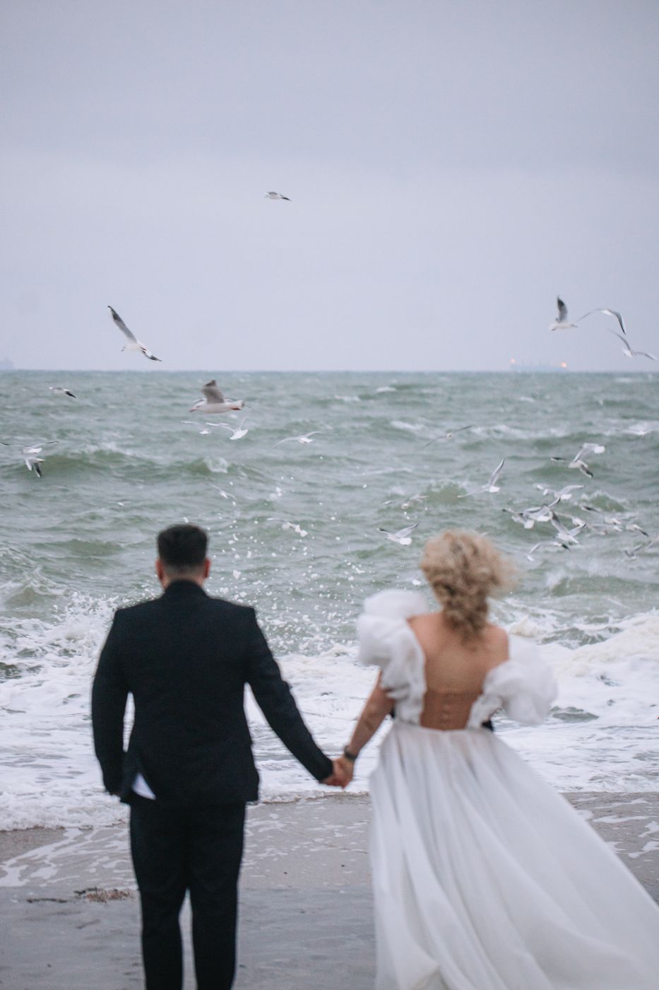A couple in wedding attire walks along a stormy beach with seagulls flying overhead.