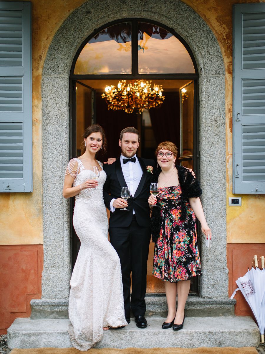 A group in formal attire gathers under stone archways at an elegant venue with chandeliers visible inside.