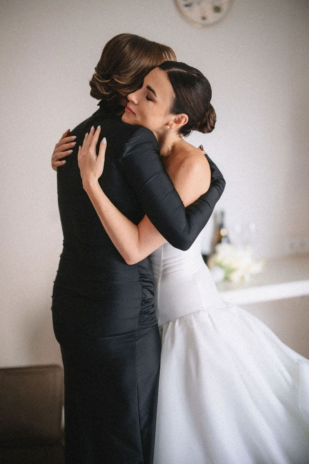 A bride in a flowing white gown embraces someone wearing black in a tender moment during a wedding celebration.