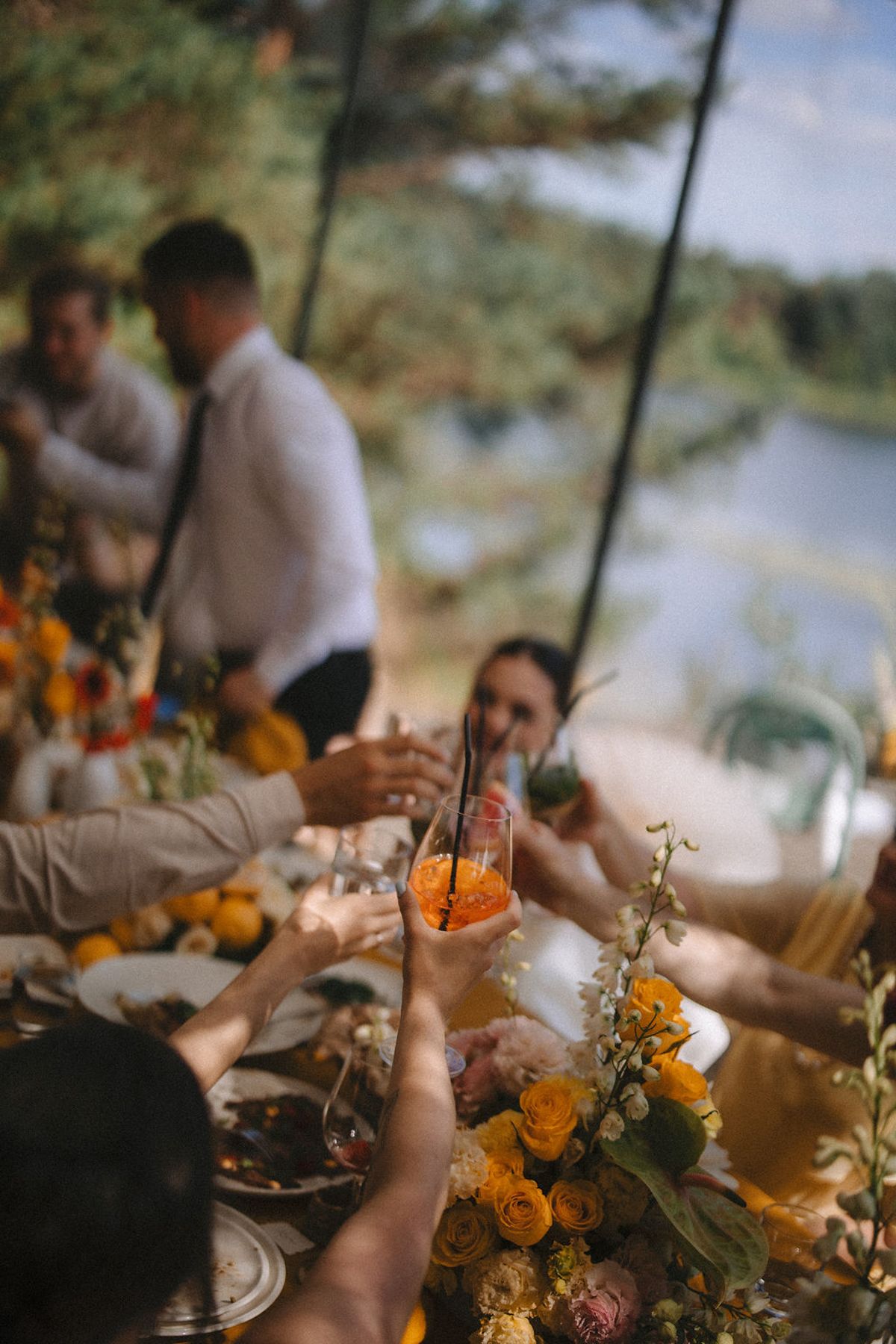 People toasting with drinks at an outdoor celebration decorated with orange flowers and table settings by a lake.