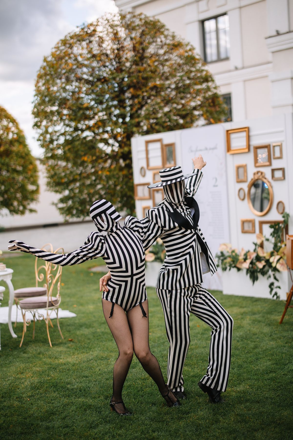 Two performers in black and white striped costumes dance playfully on a lawn in front of a white building.