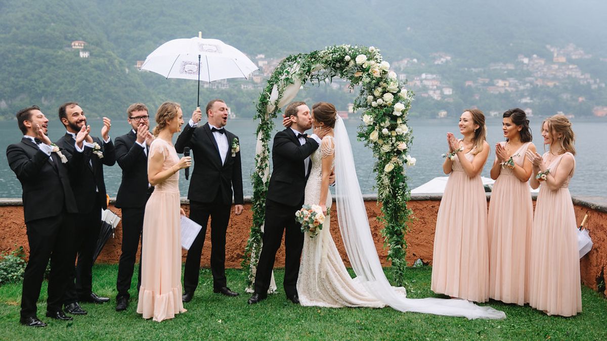 A wedding party stands on a lush green hillside overlooking a scenic mountain valley on an overcast day.
