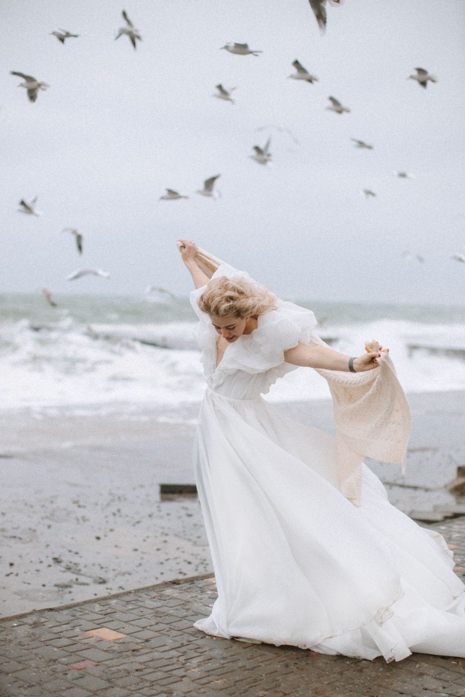 Bride in flowing dress dances on beach while seagulls fly overhead.