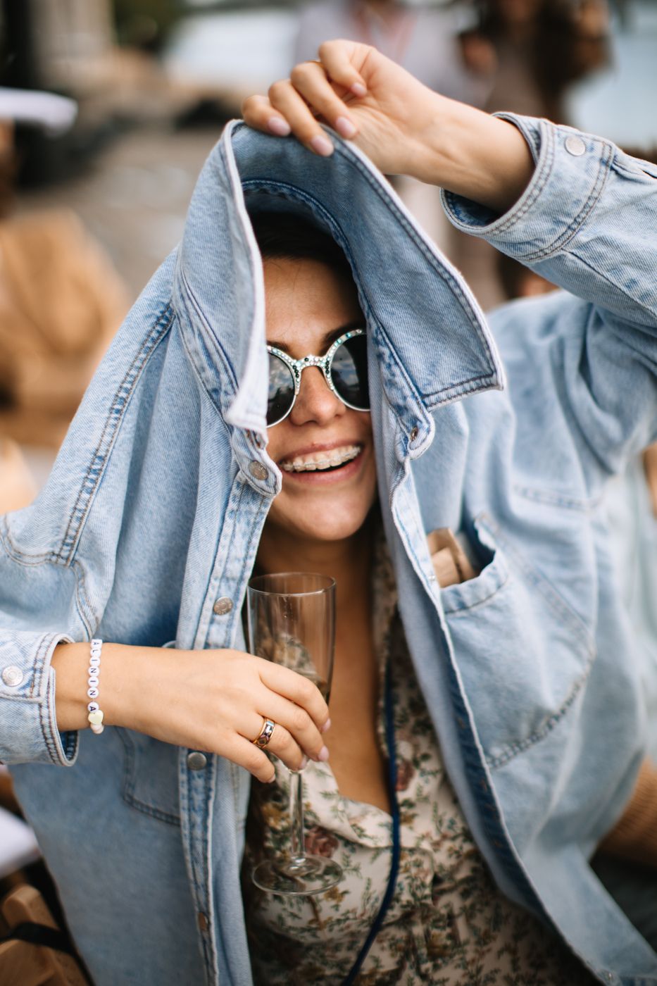 Person wearing sunglasses and denim jacket holds a drink while playfully posing with a casual, joyful expression.